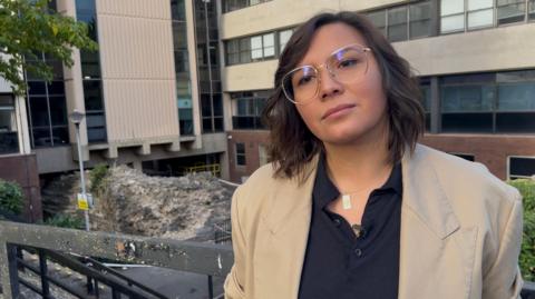 A woman with shoulder-length brown hair and large thin silver-rimmed glasses is wearing a beige blazer with a navy polo shirt. Behind and to the left of her is a Roman wall with an office building built around and above it. 
