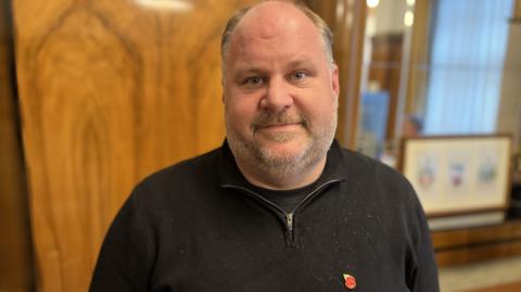 A man with a short beard and a black top stood in a room with a window and wood panels