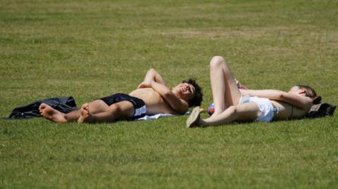 A young man and woman lying prone on grass, sunbathing. He is wearing dark shorts and she is wearing white ones. Both are flat on their backs with their left arms across their respective chests. she has her right leg raised with a bent knee.