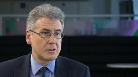 A man with grey hair and glasses in a blue suit and blue shirt against a dark background