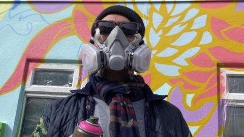 A street artist in a fume mask holding a can of pink spray paint in front of a wall spray painted with bright flowers