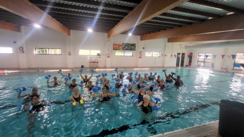 A large group of swimmers are photographed in the indoor swimming pool. The water is blue and all the swimmers are in dark-coloured swimming costumes. They are holding pool weights in the air.