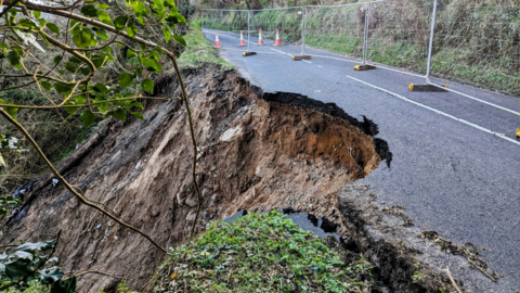 Photo of a landslide, road crumbling away into muddy slope with some vegetation in frame

The road has metal barriers and orange cones placed on it