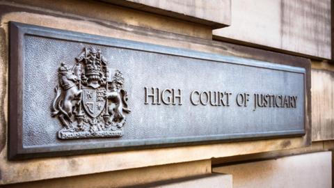 A grey metal sign on an exterior sandstone block wall of the High Court in Edinburgh. The sign has a royal crest and the words "High Court of Justiciary".