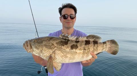 A picture of Christopher Kennedy-Barnard in a light purple t-shirt in the Middle East stood in front of the sea. He is holding a large fish with both hands and smiling at the camera. The water behind him looks calm and there is a fishing rod visible behind him. 
