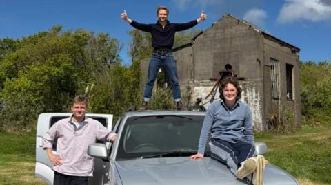 A grey car with the back door behind the drivers seat open. In front of the door is a man in a purple jumper and black trousers. He is leaning on the car. On the roof of the car is a man wearing a black jumper and blue jeans. He has both his thumbs up. 