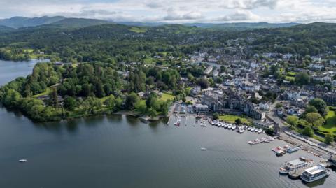 An ariel view of Windermere with the green hills of the Lake District stretching beyond its shore. Lots of boats are tied up and much of the picture is taken up with rows of houses making up a small town amid trees 