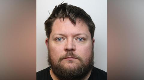 Mugshot of Chris Fox looking into the camera. He has hair and a beard and is wearing a black T-shirt