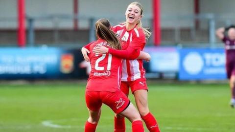 Daisy Clements celebrating a goal for Stourbridge FC, wearing a red kit with white "21" visible on the back of the shirt. There is green grass and a blurred background.