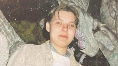 A slightly grainy photograph of Izabela Zablocka with short blonde, crew cut hair, parted in the middle. Izabela is smiling as she looks down towards the camera. She is wearing a beige denim jacket over a white knitted jumper. 