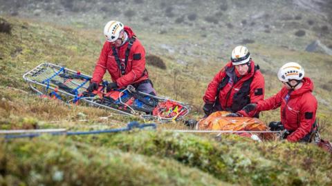 Three search and rescue team members are kneeling on a moor wearing uniform and helmets. One is working with a stretcher while the other two have large backpacks of equipment in front of them. There is a rope attached to one of them which lies in the foreground.