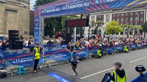 A road race finish line is set up on a city street with a large overhead banner reading “Jane Tomlinson’s Run for All.” A digital race clock suspended above the line displays 00:37:38. A runner in athletic clothing crosses the finish line with one arm raised. Event staff wearing high-visibility vests stand on either side, and a large crowd of spectators gathers behind metal barriers decorated with event branding. In the background, there are trees and multi-storey stone and brick buildings, suggesting an urban setting.