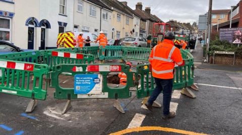 A green fence around a hole in the road with men in orange hi-vis suits in it