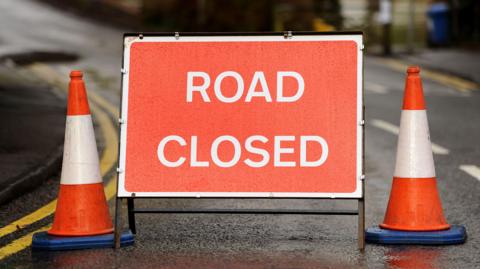 A red road closed sign with two orange cones next to it. 