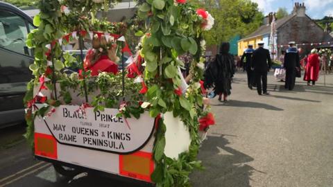 A small white carriage decorated with flowers and flags is being driven through the streets as part of the procession. The back of the vehicle says 'Black Prince Flower Boat May Queens and Kings Rame Peninsula'. People in the procession are walking in front of the vehicle. Houses are in the distance.