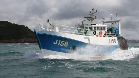 A white and blue fishing boat with J158 on the left side, in the ocean, next to another vessel out at sea with grey skies and land seen in the background.