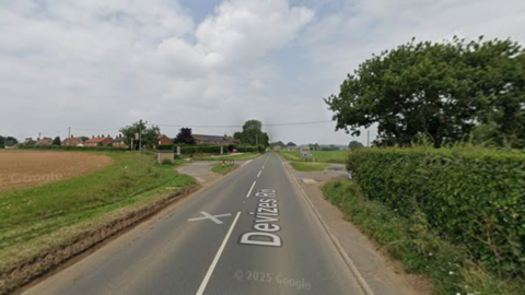 This is a Google Maps image of a road with one lane in each direction. It is a rural scene with a field on one side and a hedge on the other.