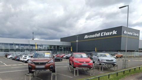 Arnold Clark headquarters in Glasgow. Cars sit in the forecourt and there is a large grey building in the background.