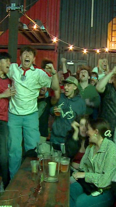 England fans celebrating as they watch the match. They are cheering and punching the air. One at the front is looking behind her at the jubilations.