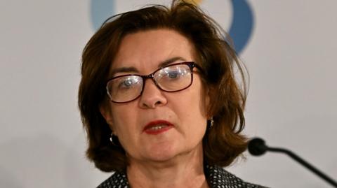  Eluned Morgan speaking at a microphone. she has dark rimmed glasses and hair, and is wearing a black jacket with white flecks in a fine pattern