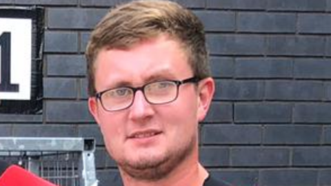 Jacob Hawden is photographed looking at the camera in front of a dark brick wall. He has short light brown hair and facial hair. He is wearing spectacles.