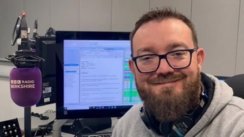 Phil Mercer, bearded and with glasses, sits in front of a computer screen in a radio studio, with a BBC Radio Berkshire branded microphone beside him