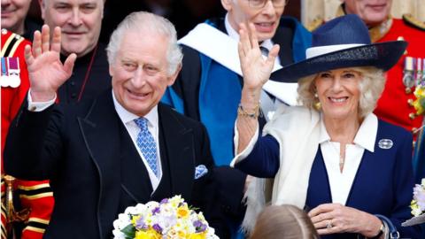 King Charles and Queen Camilla, waving, with staff behind