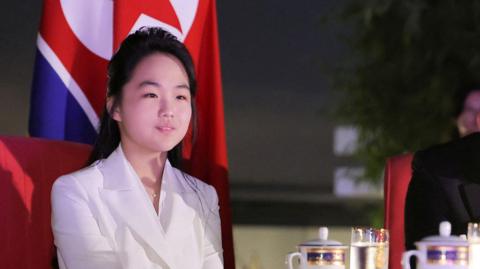 A young woman in a white suit sits at a table with the North Korean flag hung in the background 