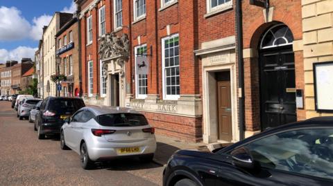 The outside of Lloyds Bank branch in Wisbech, a red-brick building with a row of cars parked outside. 