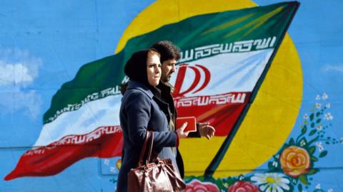 An Iranian couple walks in front of a painting of the national flag in a Tehran street