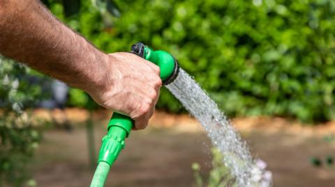 A close-up of a hand holding a green garden hose nozzle, spraying a fine stream of water downward. The background is blurred, showing green foliage and a garden.