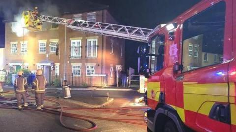 A house fire, showing a three-storey building, made from bricks, with windows, and railings outside. Two firefighters are standing outside, with brown uniforms on and helmets. A fire engine is to the right, with a crane with a firefighter in it extended towards the property. The image was taken at night.