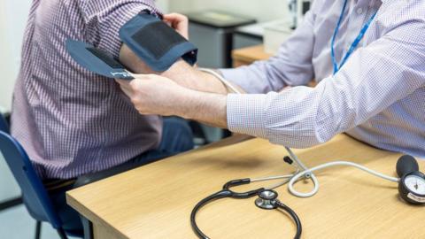 A generic image of a doctor's appointment showing a GP strapping a blood pressure monitor to a patient. Both are wearing checked shirts and sitting down. Only their torsos can be seen in the image with a desk and stethoscope in the foreground of the frame. 