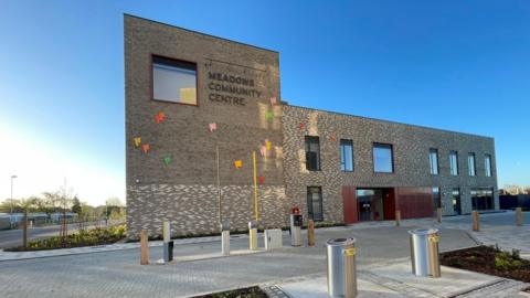 A modern brick building, which is two storeys high on the right and an extra storey high on left. It has Meadows Community Centre written on it and some colourful flags. There is a bright blue sky behind and a barrier controlled car park entrancce in front. 