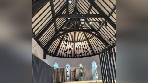 A shot of the inside of Christ Church. The image is taken from a mezzanine level and shows a vaulted ceiling with wooden beams and stained glass windows all around the higher part of the wall. The roof is more like that of a medieval banquet hall than a traditional church, and the lighting has been designed to follow a similar style. The walls are plain white.