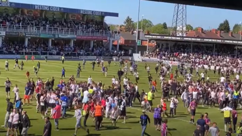 Fans of Hereford FC swarm onto the Edgar Street stadium football pitch. More than a hundred people appear to be on the pitch around the centre circle.
