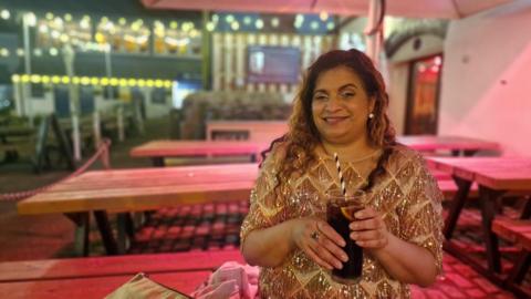 Sunita Thind sits on a bench in a gold top with a drink