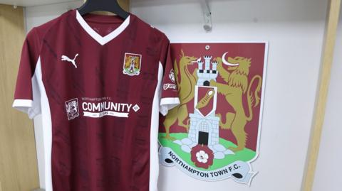 A claret and white Northampton Town home shirt hangs next the club's crest which shoes a dragon and a griffin either side of a castle with a shoe and rose. The shirt carries the name of the Northampton Town Community Trust.