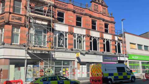 Scaffolding is on the left of the image in front of a three-storey building. Part of a police car is at the bottom on the left, near to a police van on the right.