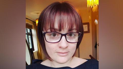 Ellie Hopkins has red and black hair and is wearing glasses. She is looking into the camera.