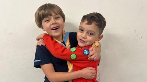 Two young boys wearing scout uniforms, smiling with their arms round each other.