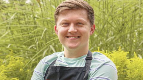 Josh Smalley, a young man with short fair hair. He is wearing a grey T-shirt under a black apron. He is looking at the camera and smiling.