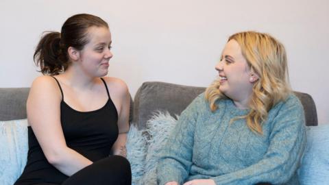 Sarah Weatherstone and Ellie facing each other on a sofa. Ms Weatherstone has dark hair tied in a pony tail and is wearing a black sleeveless top and pants. Ellie has long blonde hair and is wearing a blue-green woolly jumper.