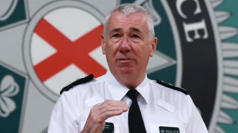 Jon Boutcher, wearing a white shirt and a black tie. There is a large PSNI logo on the wall in the background of the image. 