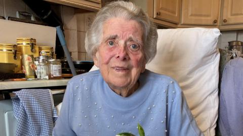 Gloria Bullen is sitting in a chair in her kitchen. She is smiling and has neatly styled hair and blue eyes. She is wearing a pale blue jumper which has pearls stitched to it. Behind her is a pillow to lean on. On the kitchen counter there are tea canisters and a jar of marmalade. 
