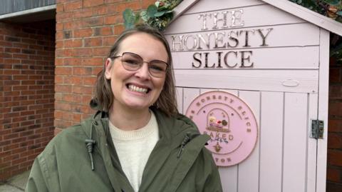 Jade Power-Vaughan, a woman wearing a green coat and a light-coloured top are standing next to a pink wooden shed which has a sign on that reads The Honest Slice.