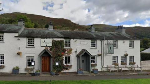 A picture of the front of the Travellers Rest Inn, near Grasmere, Cumbria. It is a converted set of traditional two-storey white terraced Georgian cottages, with the tops of some Lake District Fells in the background.