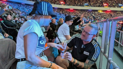 A man on one knee proposes to his partner in a packed stadium. They are both wearing Oasis T-shirts and she is wearing a bucket hat 