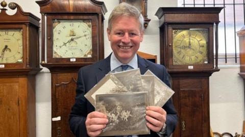 A man with grey hair and wearing a dark suit, shirt and tie holds five black and white photographs. He is standing in a white room surrounded by large wooden clocks.