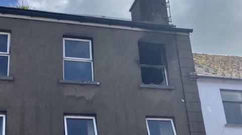 A grey building on Main Street in Ballymoney, with a chimney on top. The window on the far right is broken and there is evidence of smoke damage.